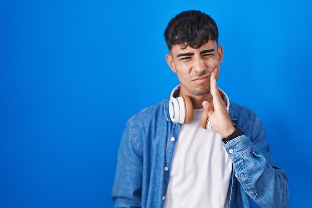 Junger hispanischer Mann, der über blauem Hintergrund steht und den Mund mit der Hand mit schmerzhaftem Ausdruck wegen Zahnschmerzen oder Zahnerkrankungen beim Zahnarzt berührt