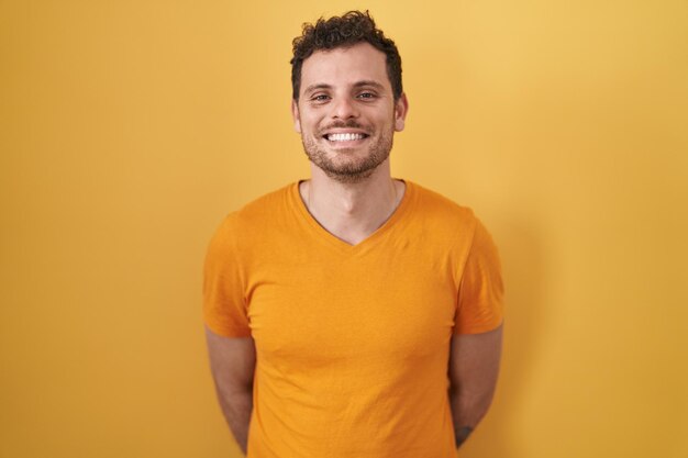 Junger hispanischer Mann, der über gelbem Hintergrund steht, mit einem glücklichen und coolen Lächeln im Gesicht, glückliche Person
