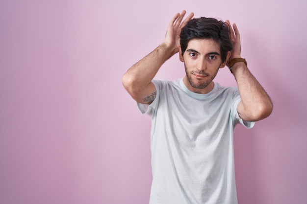 Junger hispanischer Mann, der vor rosafarbenem Hintergrund steht und Hasenohren-Gesten macht, während die Handflächen zynisch und skeptisch aussehen, zeigt das Osterhasen-Konzept