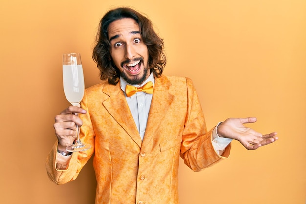 Junger hispanischer Mann im eleganten Nerd-Stil, der ein Glas prickelnden Champagner trinkt und den Erfolg mit einem glücklichen Lächeln und Siegerausdruck mit erhobener Hand feiert