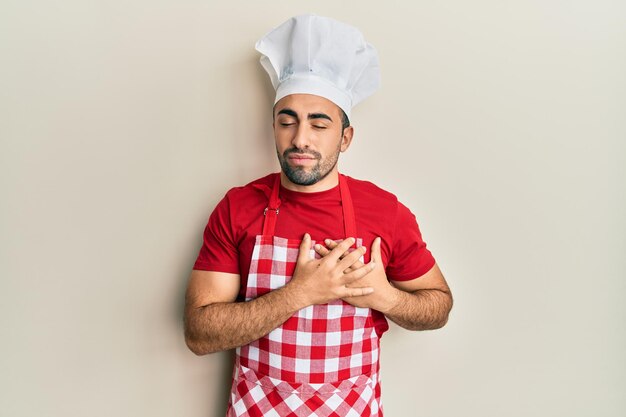 Junger hispanischer Mann in Bäckeruniform, lächelnd mit den Händen auf der Brust, geschlossenen Augen und dankbarer Geste im Gesicht, Gesundheitskonzept