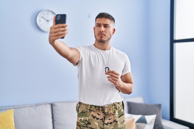 Junger hispanischer Mann in Camouflage-Armeeuniform macht Selfie zu Hause skeptisch und nervös, runzelt die Stirn wegen des Problems. negativer Mensch.