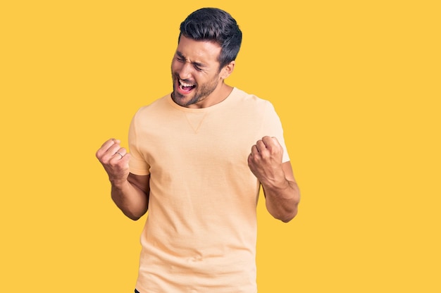 Junger hispanischer Mann in lässiger Kleidung freut sich über den Erfolg mit erhobenen Armen und geschlossenen Augen und feiert das Konzept des lächelnden Gewinners des Sieges