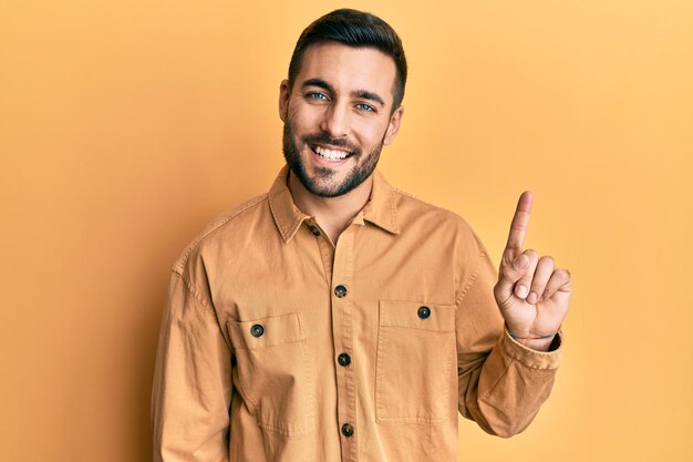 Junger hispanischer Mann in lässiger Kleidung, lächelnd mit einer Idee oder Frage, zeigend mit dem Finger nach oben und einem glücklichen Gesicht Nummer eins