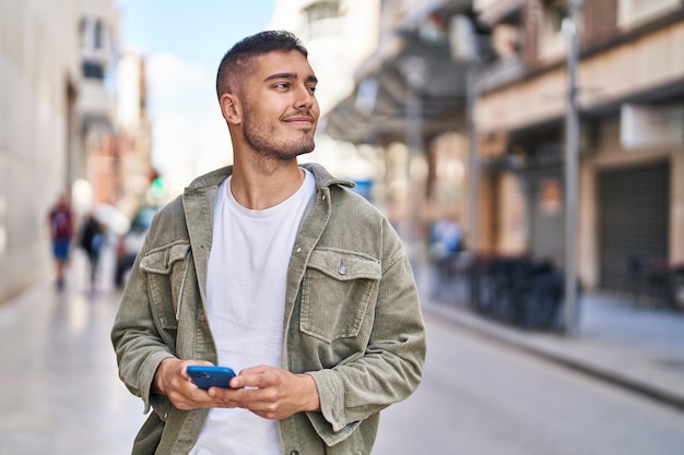 Junger hispanischer Mann lächelt selbstbewusst mit Smartphone auf der Straße