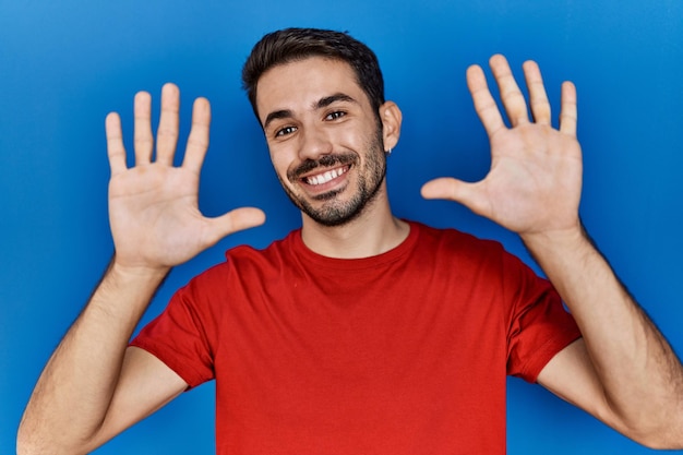 Junger hispanischer Mann mit Bart, der ein rotes T-Shirt über blauem Hintergrund trägt und mit den Fingern Nummer zehn nach oben zeigt, während er selbstbewusst und glücklich lächelt.