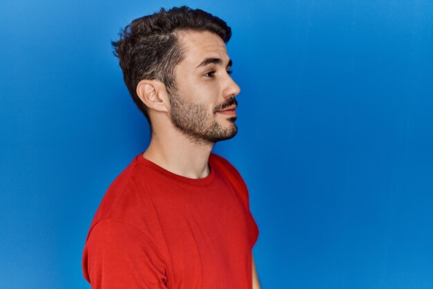 Junger hispanischer Mann mit Bart, der ein rotes T-Shirt über blauem Hintergrund trägt und zur Seite schaut, entspannte Profilpose mit natürlichem Gesicht und selbstbewusstem Lächeln.