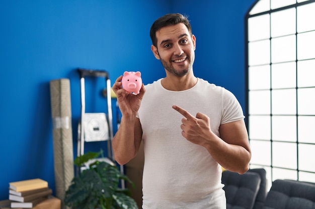 Kostenloses Foto junger hispanischer mann mit bart, der ein sparschwein im neuen zuhause hält, lächelt glücklich und zeigt mit hand und finger