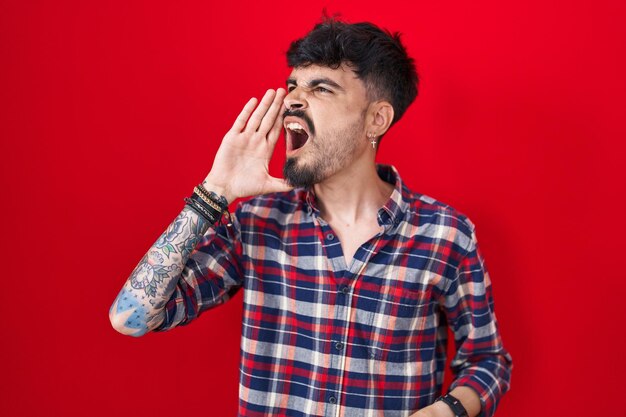 Junger hispanischer Mann mit Bart, der über rotem Hintergrund steht und laut schreit und mit der Hand auf dem Mund Kommunikationskonzept zur Seite schreit