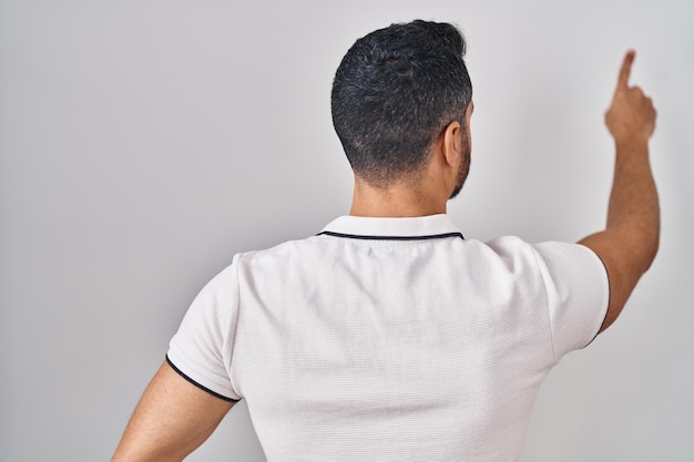 Kostenloses Foto junger hispanischer mann mit bart in lässiger kleidung vor weißem hintergrund posiert rückwärts und zeigt mit der fingerhand nach vorne