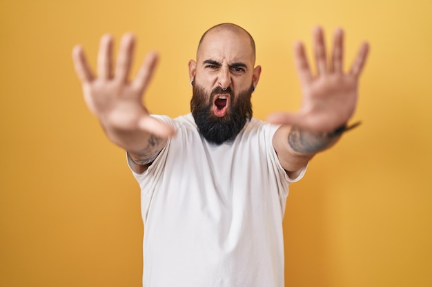 Kostenloses Foto junger hispanischer mann mit bart und tätowierungen steht vor gelbem hintergrund und macht stopp-geste mit wütendem und frustriertem ausdruck an den handflächen