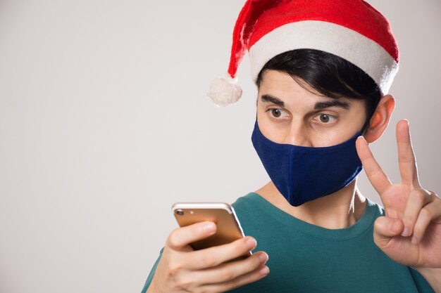 Junger hispanischer Mann mit einer Gesichtsmaske und einer Weihnachtsmütze, die auf sein Telefon schauen und eine Friedensgeste tun