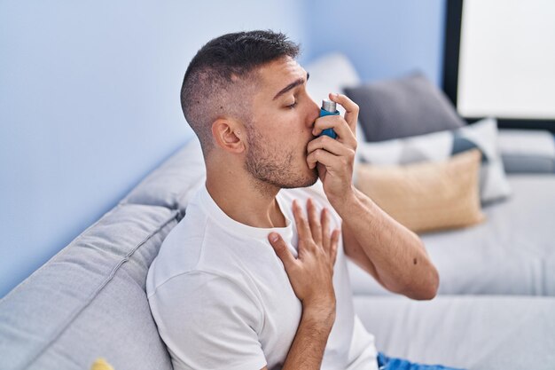 Junger hispanischer Mann mit Inhalator, der zu Hause auf dem Sofa sitzt