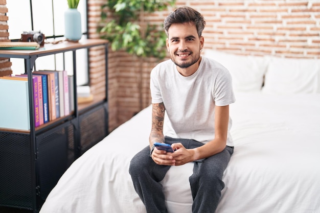 Junger hispanischer Mann mit Smartphone sitzt im Schlafzimmer auf dem Bett