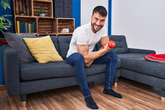 Junger hispanischer Mann trainiert mit Hanteln, die zu Hause auf dem Sofa sitzen