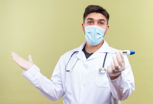 Junger hübscher Arzt, der weiße medizinische Handschuhe der blauen medizinischen Maske weißes medizinisches Kleid weiße medizinische Handschuhe und Stethoskop hält, die elektronisches Thermometer halten, das über khaki Wand steht