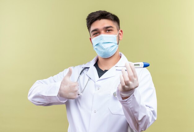 Junger hübscher Arzt, der weiße medizinische Handschuhe der blauen medizinischen Maske weißes medizinisches Kleid weiße medizinische Handschuhe und Stethoskop hält, die elektronisches Thermometer halten, glückliche Daumen hoch stehend über khaki backgrou tun