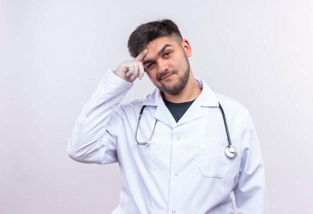 Junger hübscher Arzt, der weiße medizinische Handschuhe des weißen medizinischen Kleides und das Stethoskop trägt, das nachdenklich über der weißen Wand stehend schaut