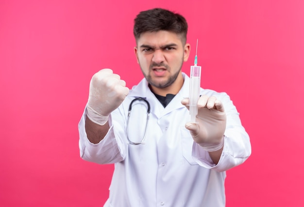 Junger hübscher Arzt, der weiße medizinische Handschuhe des weißen medizinischen Kleides und Stethoskop trägt, das mit einer Injektion droht, die über rosa Wand steht