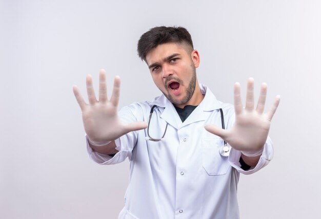 Junger hübscher Arzt, der weiße medizinische Handschuhe des weißen medizinischen Kleides und Stethoskop trägt, das Stoppschild mit den Händen steht, die über weißer Wand stehen