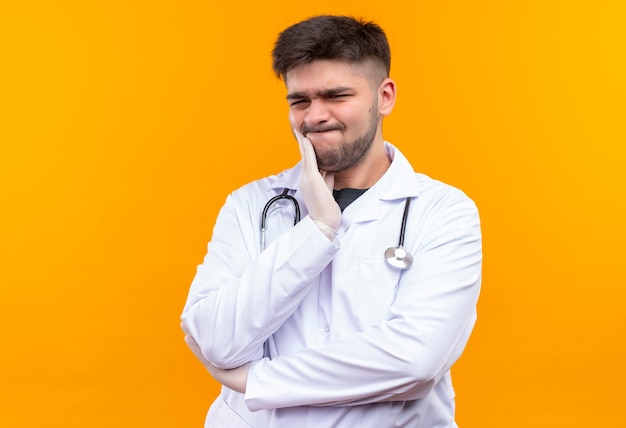 Junger hübscher Arzt, der weiße medizinische Handschuhe des weißen medizinischen Kleides und Stethoskop trägt, das unter Zahnschmerzen leidet, die über orange Wand stehen