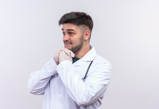 Junger hübscher Arzt, der weiße medizinische Handschuhe des weißen medizinischen Kleides und Stethoskop trägt, die neben fasziniertem Stehen über weißer Wand suchen