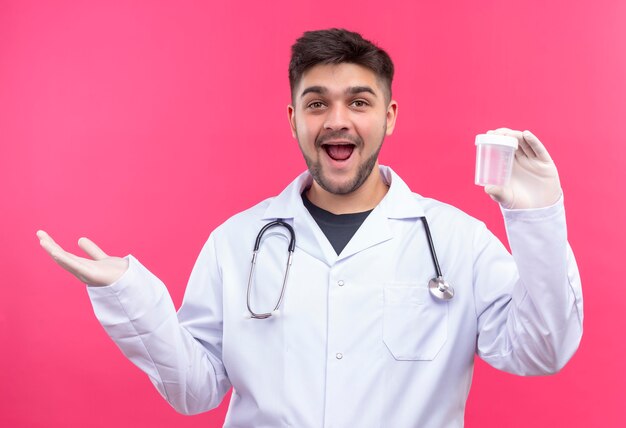 Junger hübscher Arzt, der weiße medizinische Handschuhe des weißen medizinischen Kleides und Stethoskop trägt, froh für Ergebnisse in transparentem Analysebehälter, der über rosa Wand steht