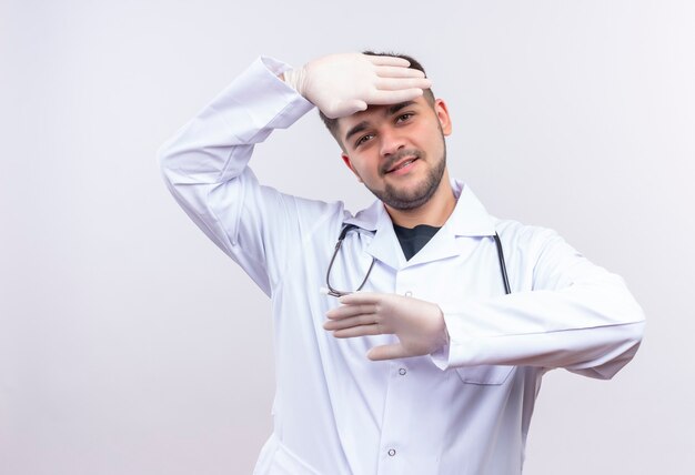 Junger hübscher Arzt, der weiße medizinische Handschuhe und Stethoskop des weißen medizinischen Kleides trägt und mit den Händen spielt, die über weißer Wand stehen