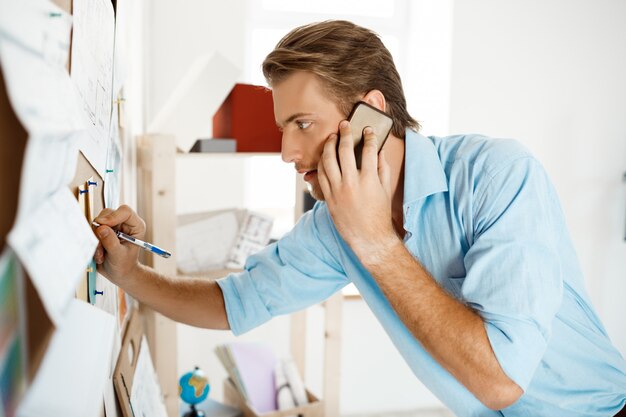 Junger hübscher Geschäftsmann, der an dem Papier schreibt, das an die Pinnwand geheftet wird und am Telefon spricht.