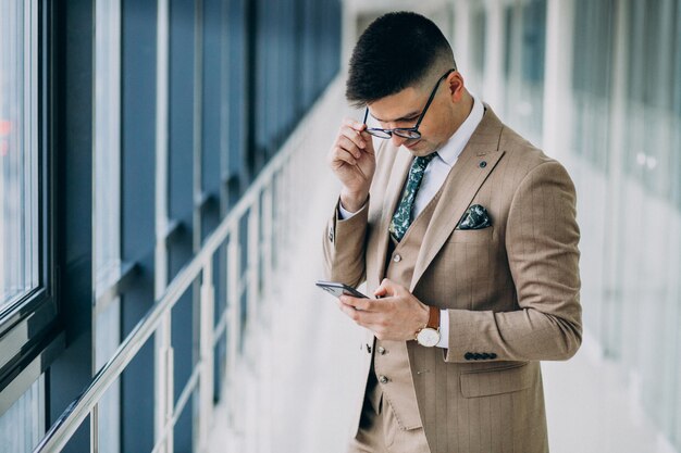 Junger hübscher Geschäftsmann, der mit Telefon im Büro steht