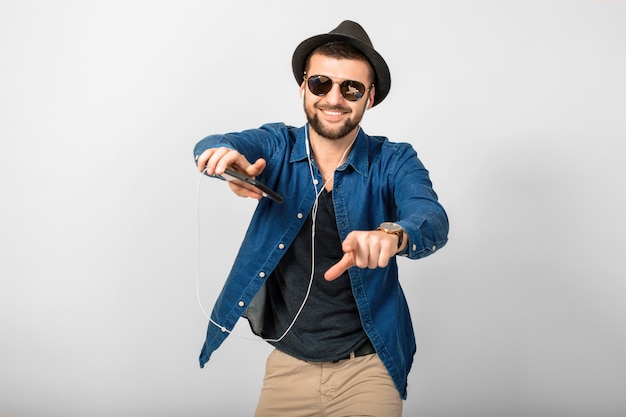 Junger hübscher glücklicher lächelnder Mann, der Musik in den Kopfhörern lokalisiert auf weißem Studiohintergrund hört, Smartphone hält, Jeanshemd, Hut und Sonnenbrille tragend