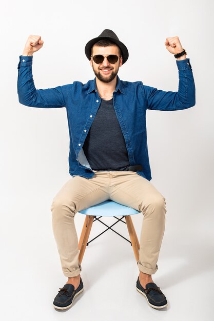 Junger hübscher Hipster-Mann, isolierter weißer Studiohintergrund, stilvolles Outfit, Jeanshemd, Hose, Hut, Sonnenbrille, auf Stuhl sitzend, Erfolg, Gewinn, emotional, glücklich, lächelnd