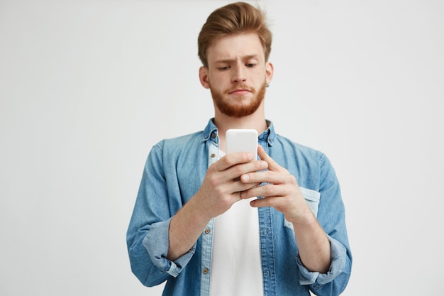 Junger hübscher Kerl, der die Stirn runzelt und den Telefonbildschirm beim Surfen im Internet betrachtet.