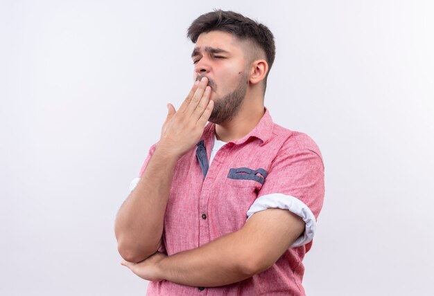 Junger hübscher Kerl, der rosa Poloshirt gähnt, das über weißer Wand gähnt