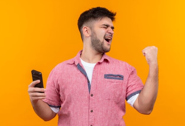 Junger hübscher Kerl, der rosa Poloshirt hält, das Telefon glücklich macht, tat es mit Faust, die über orange Wand steht