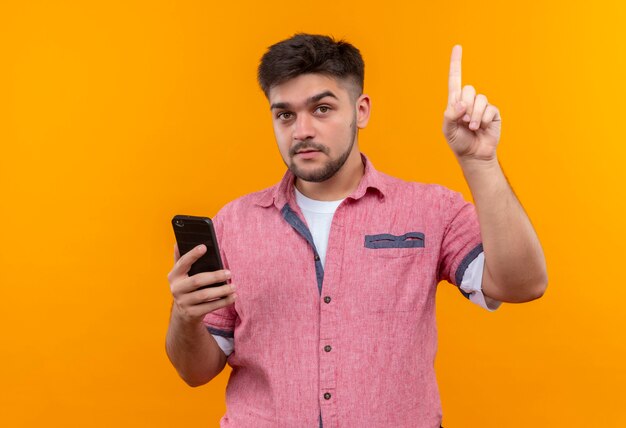 Junger hübscher Kerl, der rosa Poloshirt hält, das Telefon konzentriertes Zeigen zeigt, wobei Zeigefinger über orange Wand steht