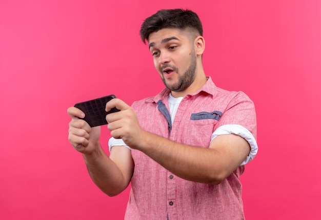 Junger hübscher Kerl, der rosa Poloshirt spielt Spiel am Telefon steht über rosa Wand