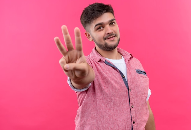 Junger hübscher Kerl, der rosa Poloshirt trägt, das drei Zeichen mit den Fingern steht über rosa Wand zeigt