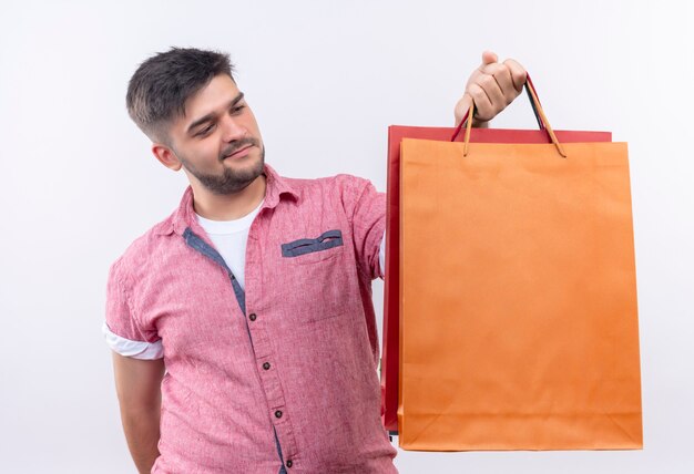 Junger hübscher Kerl, der rosa Poloshirt trägt, das Einkaufstaschen hält, die über weißer Wand stehen