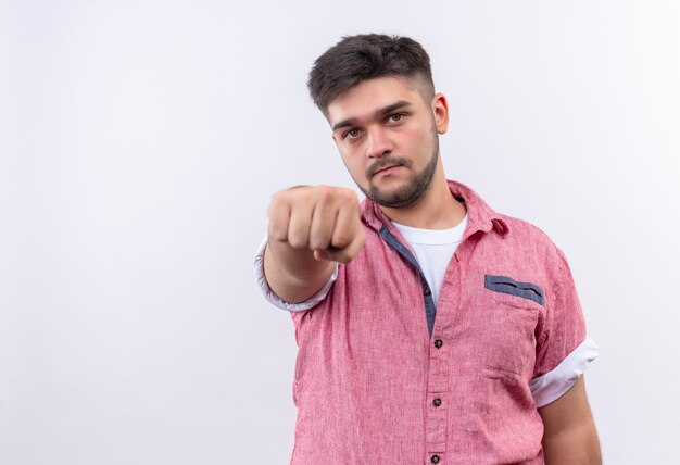 Junger hübscher Kerl, der rosa Poloshirt trägt, das ernsthaft Faust zeigt, die über weißer Wand steht