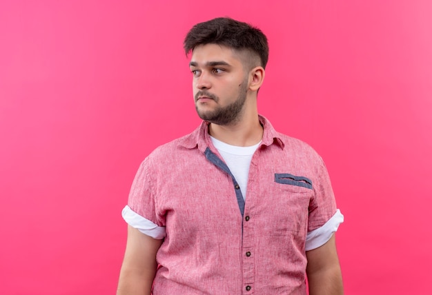 Junger hübscher Kerl, der rosa Poloshirt trägt, das ernsthaft neben dem Stehen über rosa Wand schaut