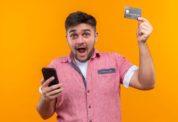 Junger hübscher Kerl, der rosa Poloshirt trägt, das glücklich hält Telefon hält, das Kreditkarte über orange Wand steht