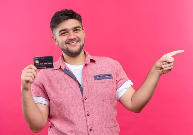 Junger hübscher Kerl, der rosa Poloshirt trägt, das glücklich Kreditkarte hält, die nach links mit Zeigefinger zeigt, der über rosa Wand steht