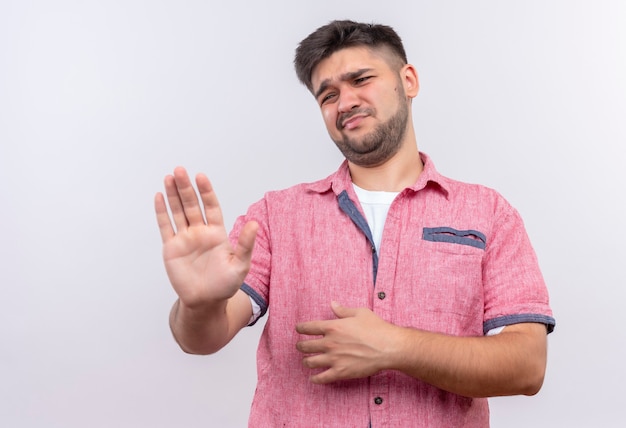 Junger hübscher Kerl, der rosa Poloshirt trägt, das gut gefüttertes Zeichen mit Armen tut, die über weißer Wand stehen