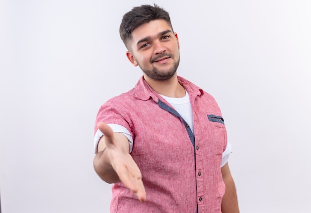 Junger hübscher Kerl, der rosa Poloshirt trägt, das koridal Hand zum Händeschütteln gibt, das über weißer Wand steht