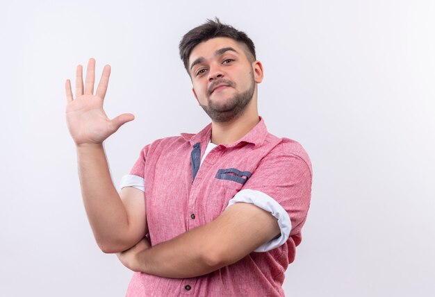 Junger hübscher Kerl, der rosa Poloshirt trägt, das lächelt und fünf mit den Fingern zeigt, die über weißer Wand stehen