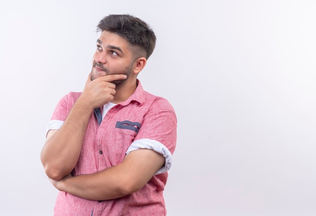 Junger hübscher Kerl, der rosa Poloshirt trägt, das nachdenklich oben schaut, außer oben über weißer Wand