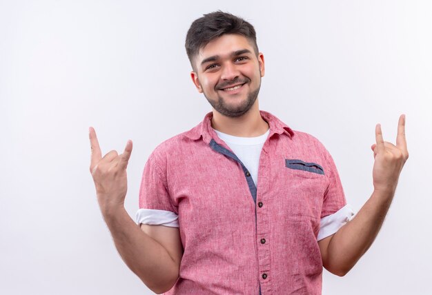 Junger hübscher Kerl, der rosa Poloshirt trägt, das Rock für immer mit Fingern unterzeichnet, die glücklich über weißer Wand stehen