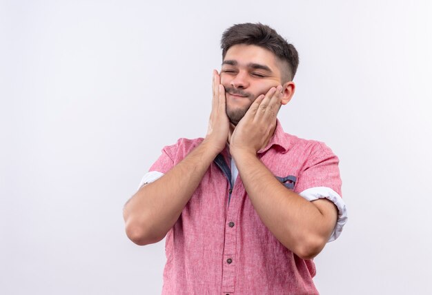 Junger hübscher Kerl, der rosa Poloshirt trägt, das sein Gesicht massiert, das über weißer Wand steht