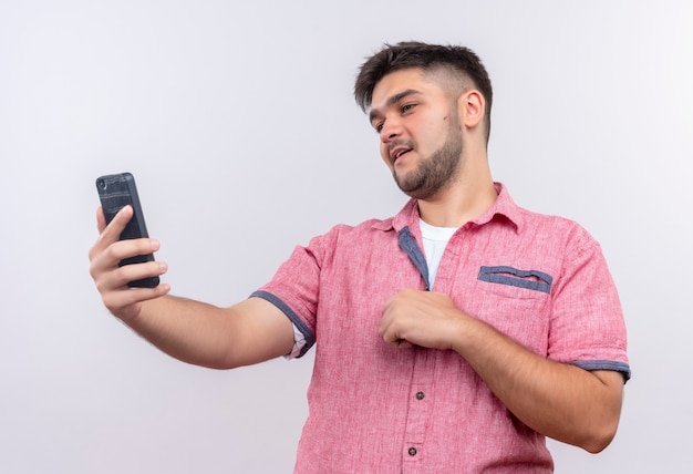 Junger hübscher Kerl, der rosa Poloshirt trägt, das Selfie erfreut macht, über weißer Wand zu stehen